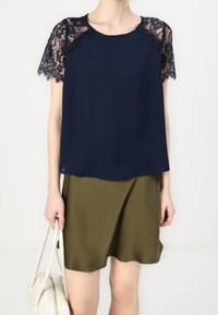 Kvinna i marinblå topp med spetsärmar och olivgrön kjol, som håller en vit handväska med båda händerna mot en neutral bakgrund.