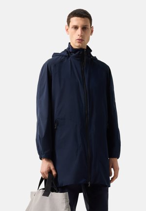 Mand iført en navy blå hættetrøje med lynlås, der holder en lysegrå tote taske med sorte håndtag mod en ensfarvet baggrund.