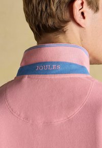 Rozā polo krekls ar zilu apkakli, ar izšūtu "JOULES" logo. Tekstūrēta auduma un kontrastējošas šuve detaļas.