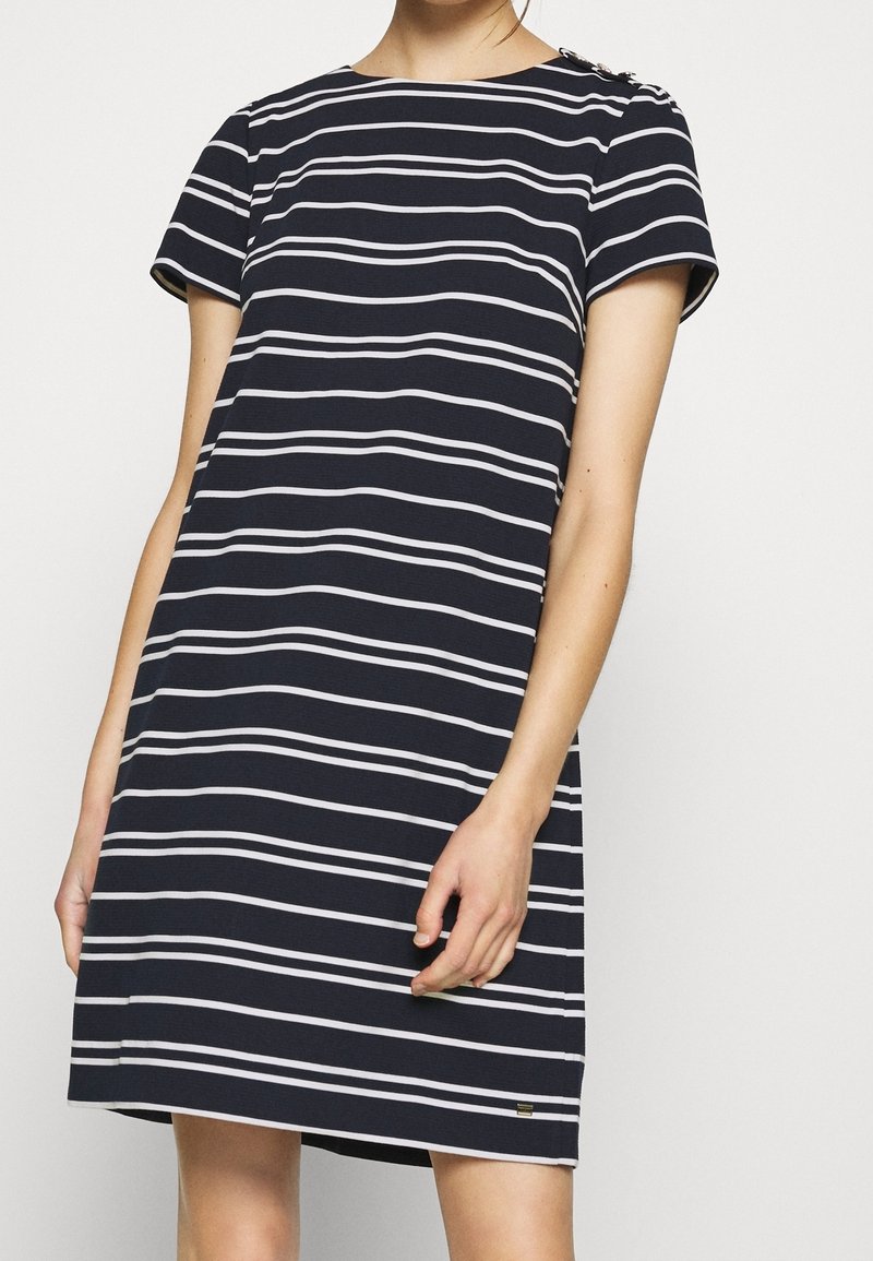 Femme portant une robe bleue marine à manches courtes avec des rayures blanches horizontales et un détail de boutons sur l'épaule, debout devant un fond uni.