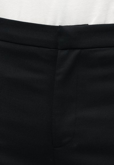 Κοντινό πλάνο ενός ατόμου που φοράει μαύρο παντελόνι με επίπεδη εμπρόσθια όψη και ένα λευκό πουκάμισο που είναι μπαίνει μέσα στο παντελόνι.