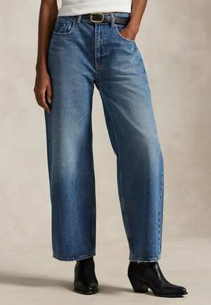 Modèle portant un jean bleu à jambes larges, une ceinture noire et des bottines noires ; une main repose dans une poche.
