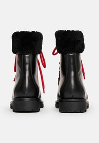 Botas de invierno de cuero negro con gruesos puños de piel negra y cordones rojos, mostradas desde atrás sobre un fondo blanco.