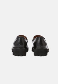 Paire de mocassins en cuir noir avec semelles épaisses à relief, vus de dos sur un fond clair uni.