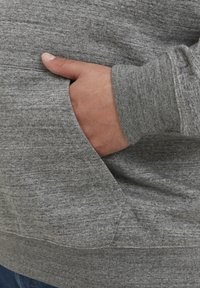 Grauer Pullover aus strukturiertem Stoff mit einer Seitentasche und gerippten Bündchen. Nahaufnahme einer Hand, die in der Tasche ruht.