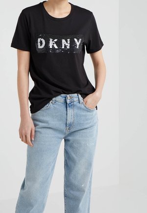Camiseta negra de manga corta con un logo DKNY de lentejuelas en blanco, combinada con jeans de tiro alto en azul claro. La tela es suave y cómoda.