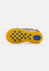 Krāsaina sporta apavu zole ar dzeltenu rakstu, kas rotāta ar zilām apaļām saķeres akcentiem un teksturētu apdari, lai nodrošinātu saķeri. Redzams zīmola logotips.