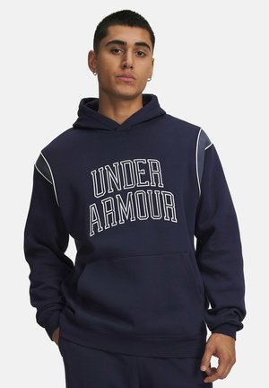 Jonge man die een marineblauwe hoodie draagt met de tekst "UNDER ARMOUR" in witte blokletters, handen in de zakken en kleine oorringen.