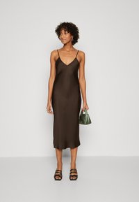 Robe de soirée en satin marron avec des fines bretelles, longueur genoux. Le modèle tient un sac vert et porte des sandales à lanières noires. Texture lisse, design minimal.