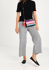 Farverig crossbody taske med zigzag mønster i pink, blå og sort. Har frynser og en lang justerbar rem. Kombineret med stribede bukser.