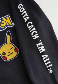 Juodas audinys, ant kurio siuvinėtas geltonas Pikachu veidas su raudonomis skruostų dėmėmis ir mirksinčia akis. Ant rankovės baltais rašmenimis parašyta "REIKIA SURINKTI VISUS!™."