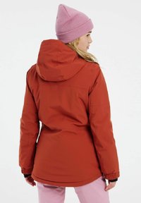 Rote isolierte Jacke mit Kapuze, elastischen Bündchen und abgerundetem Saum. Das Model trägt eine hellrosa Mütze und hellrosa Hosen.