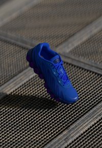 Blauwe sportieve schoen met paarse zoolaccenten op een metalen roosteroppervlak in fel buitenlicht.