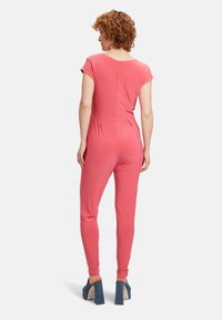 Lyserød jumpsuit med korte ærmer, tætsiddende design, lynlås på ryggen og sidelommer. Båret med denimplatformsandaler.
