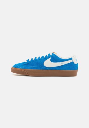 Niebieskie zamszowe sneakersy z białym logo Nike Swoosh, gumową brązową podeszwą i białymi sznurowadłami. Charakteryzują się detalami przeszycia oraz wyściełanym kołnierzem dla komfortu.