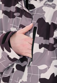 Takki, jossa on harmaan ja violetin sävyissä oleva camouflagemalli, jossa on vetoketjullinen tasku ja säädettävät hihansuut. Valmistettu sileästä synteettisestä materiaalista.