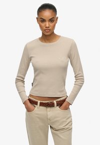 Lysebeige langærmet top med rund halsudskæring og ribbet tekstur, der har diskret branding. Kombineret med et brunt vævet bælte over lyse bukser.