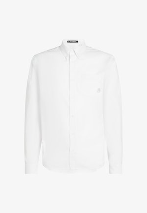 Valkoinen pitkähihaista paitaa, joka on valmistettu puuvillasta. Paitassa on napillinen kaulus, rintatasku ja hienovarainen logopistotus taskussa.