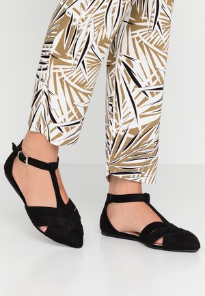Pieds portant des sandales plates noires à bride en T avec boucle, accompagnés d'un pantalon court à motif marron et blanc, sur fond blanc.