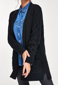 Černý pletený cardigan s texturovanými kabelkovými vzory, dlouhými rukávy, otevřeným předním dílem a postranními rozparky, nošený přes modrou džínsovou košili.