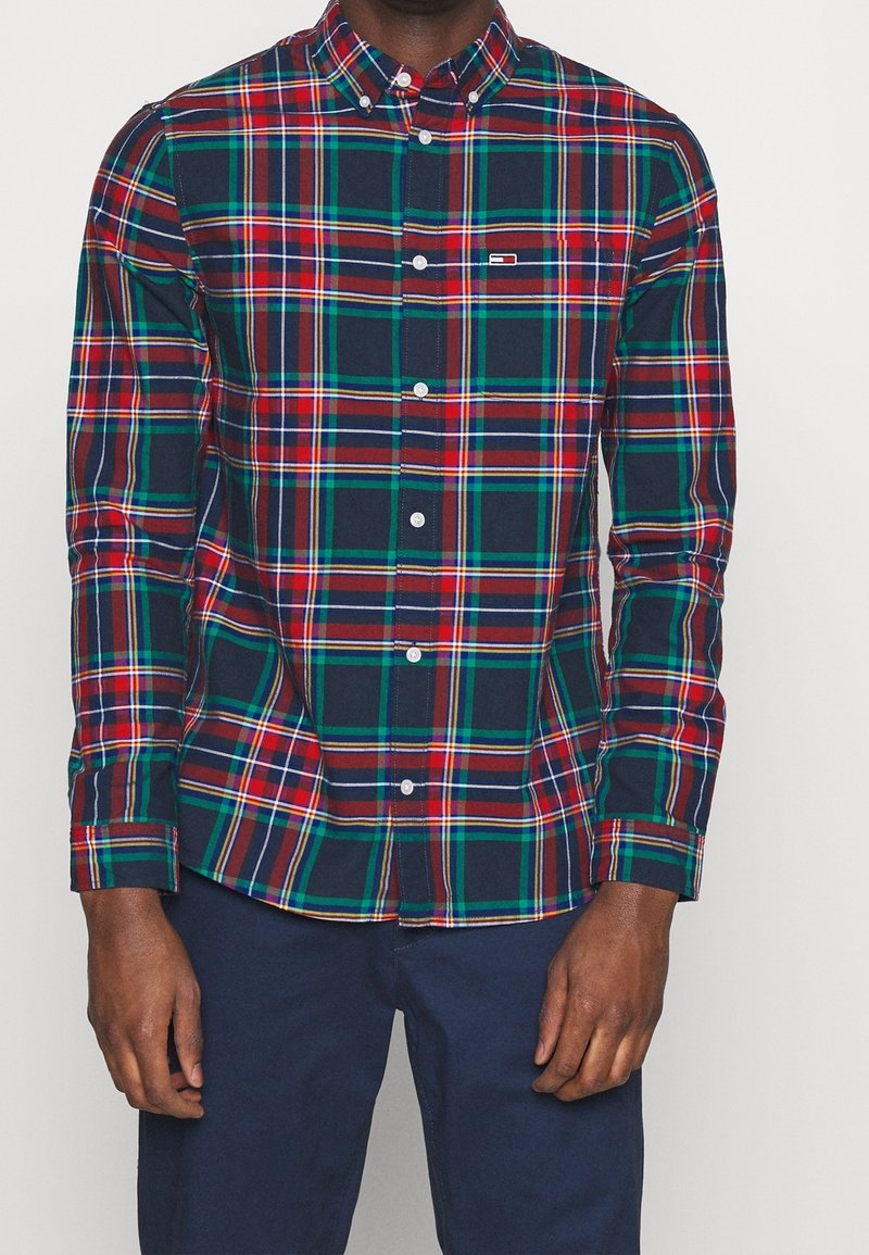 Homme portant une chemise à manches longues boutonnée, à motif écossais rouge, vert et bleu marine, et un pantalon bleu marine, faisant face à l'avant.