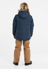 Giubbotto imbottito blu con cappuccio, polsini elasticizzati e etichetta 'PROTEST' sul colletto, abbinato a pantaloni da neve beige e stivali neri. Tessuto liscio.