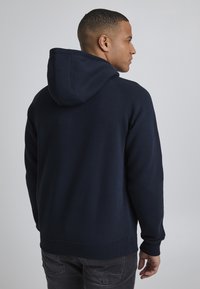 Tamsiai mėlyna džemperis su gobtuvu, pagamintas iš medvilnės mišinio, su priekinėmis kišenėmis, įsiūtomis rankovėmis ir pirkimo džiunglėm. Lygus paviršius, kasdieniškas dizainas.