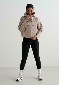 Donna sorridente che si chiude una giacca beige con cappuccio, indossa leggings neri, calzini bianchi e sneakers grigio-bianco, in piedi contro un muro bianco semplice.