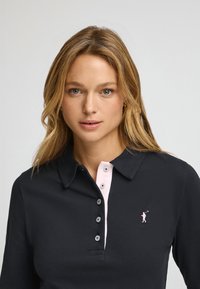 Camisa polo negra con una tapeta rosa, que presenta cinco botones grises y un pequeño logo rosa de un golfista en el lado izquierdo.