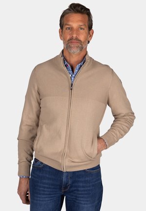 Beige zip-up cardigan with ribbed texture at shoulders, knitted fabric, stand collar, and side pockets, worn over a patterned shirt.