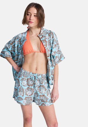 Młoda kobieta ubrana w pomarańczowy top od bikini z niebieską i brązową kwiecistą koszulą oraz pasujące szorty, stoi z jedną ręką na biodrze, patrząc w lewo.