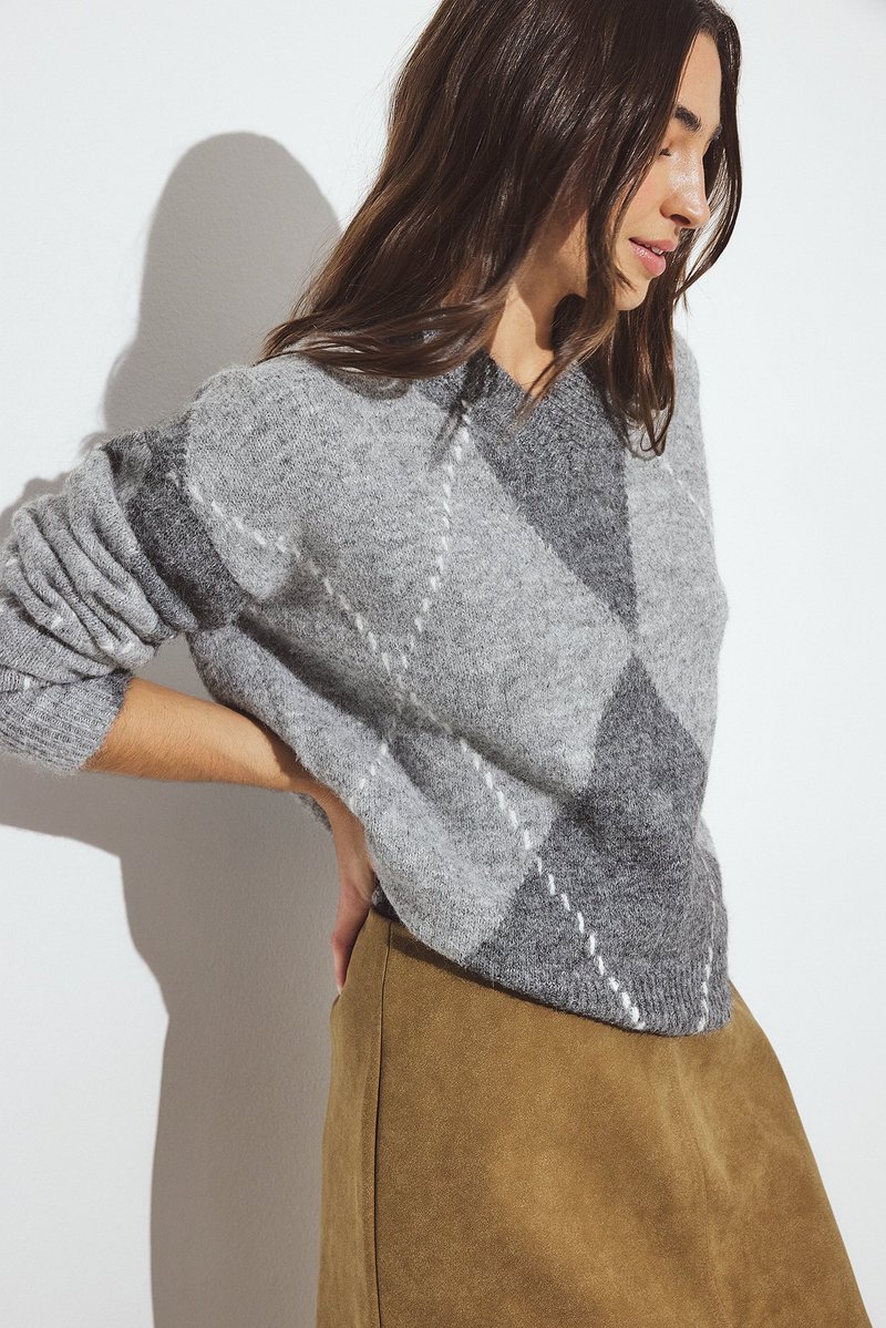 Siv š sweater z vzorcem diamanti, mehak na dotik in zavihki na rokavih, v kombinaciji z rjavim suknjičem iz usnja. Minimalistična ozadje.
