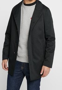 Cappotto nero con ampio colletto, bottoni doppi e una texture liscia, indossato sopra una felpa grigia e jeans blu.
