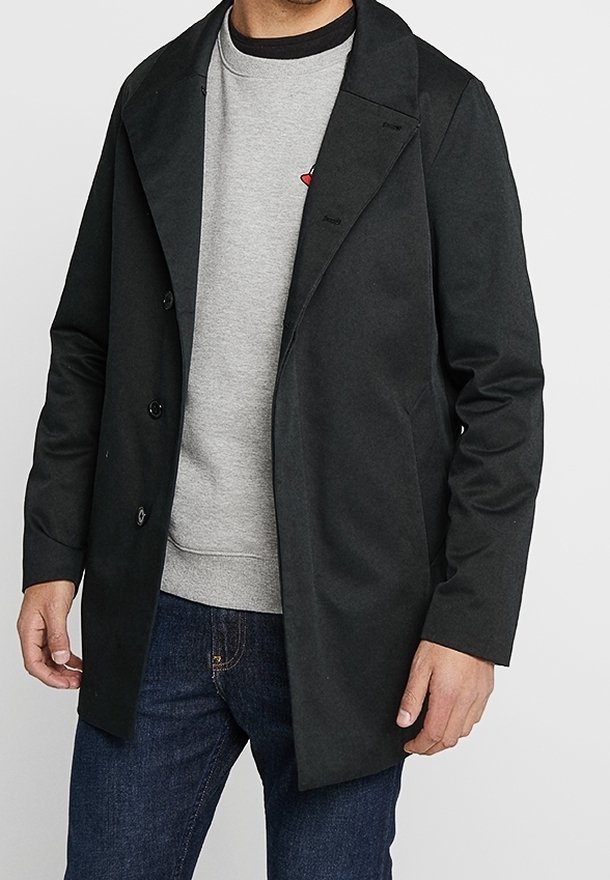 Cappotto nero con ampio colletto, bottoni doppi e una texture liscia, indossato sopra una felpa grigia e jeans blu.