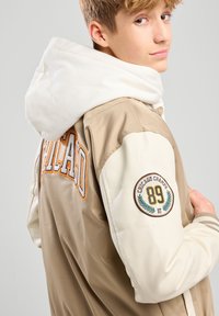 Beige ja cream bomber takki, kus on "CHICAGO" tikand, ümmargune plekk varrukal ja pehme valge kapuuts. Tekstuuriga kangas.