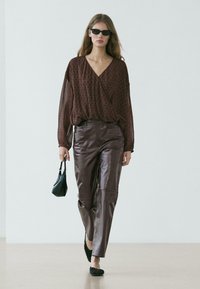 Frau geht, trägt eine braune transparente Bluse, dunkelbraune Lederhose, schwarze Ballerinas, Sonnenbrille und hält eine kleine schwarze Handtasche.