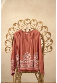 Rostfarbige Bluse mit langen Ärmeln, V-Ausschnitt und weißer Blumenstickerei am Saum und an den Ärmeln, präsentiert auf einem dekorativen Rattanstuhl.