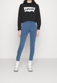 Svart croppad hoodie med en silver Levi's-logotyp, i kombination med högmidjade blå skinny jeans och vita högtopps sneakers med svarta ränder.