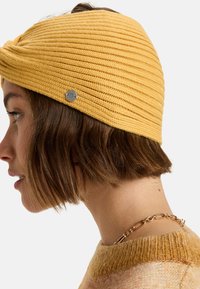 Gele gebreide hoofdband met een geprofileerd ribbelontwerp, voorzien van een kleine metalen accent. Korte bruine haren zijn zichtbaar, met een gouden kettingketting.