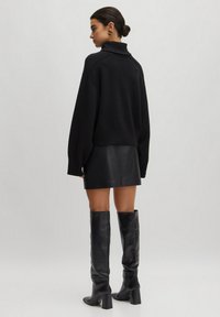 Zwarte gebreide trui met een hoge kraag, gecombineerd met een zwarte leren minirok en zwarte kniehoogte laarzen met hak. Glad textuur en aangesloten ontwerp.