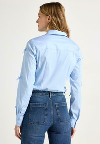 Camicia di cotone azzurro con maniche lunghe e fiocchi ai polsini, abbinata a jeans in denim scuro a vita alta.