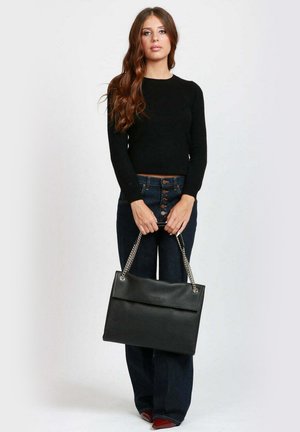 Borsa a tracolla nera in pelle con finitura texturizzata, dotata di patta superiore e manico a catena, tenuta da una persona in jeans scuri e una maglia nera.