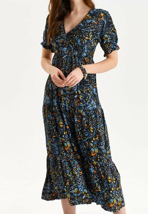 Kobieta w czarnej sukience midi z niebieskimi, pomarańczowymi i żółtymi kwiatowymi wzorami, krótkimi bufiastymi rękawami i dekoltem w kształcie litery V, nosząca ciemne bransoletki z koralików.