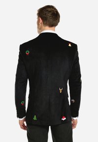 Man med svart blazer dekorerad med små broderade julikoner inklusive kransar, renar, klockor, träd, tomteluva och julpudding.