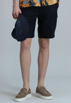 Shorts navy, mocassins beige en suede avec des semelles blanches, et un chapeau seau bleu foncé. Chemise à manches courtes à motifs floraux avec des tons orange et bleu.