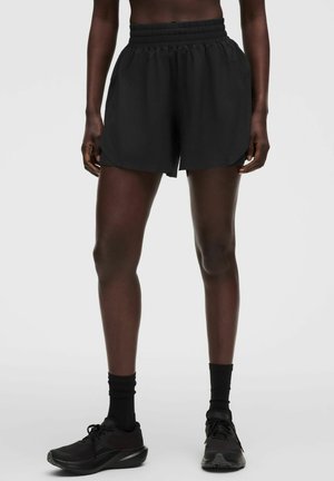 Athlet trägt schwarze Shorts mit elastischem Bund, schwarze Knöchelsocken und schwarze Laufschuhe und steht vor einem schlichten Hintergrund.