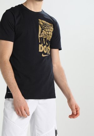 Man draagt een zwart Nike "Just Do It" t-shirt met een gouden basketbalring-graphic en witte korte broek tegen een witte achtergrond.