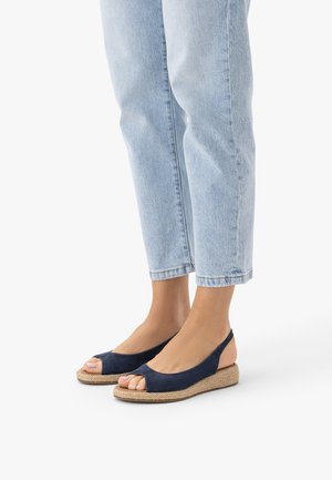 Stopy w granatowych koturnach na otwartym palcu z paskiem z tyłu, z plecioną podeszwą, w połączeniu z jasnoniebieskimi dżinsami o skróconej długości na białym tle.