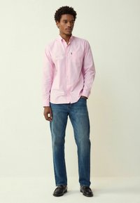 Cămașă roz cu dungi, cu nasturi și un buzunar pe piept, asortată cu blugi albaștri și mocasini negri, stând pe un fundal simplu.