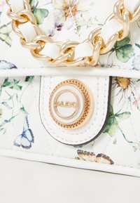 Sac à main blanc à imprimé floral avec une chaîne dorée et un détail de logo circulaire sur la fermeture magnétique. Fini en cuir texturé.